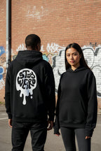 Two people wearing black hoodies with a logo on a graffiti-covered wall background