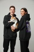 Two people wearing black hoodies, one holding a cat, on a plain background