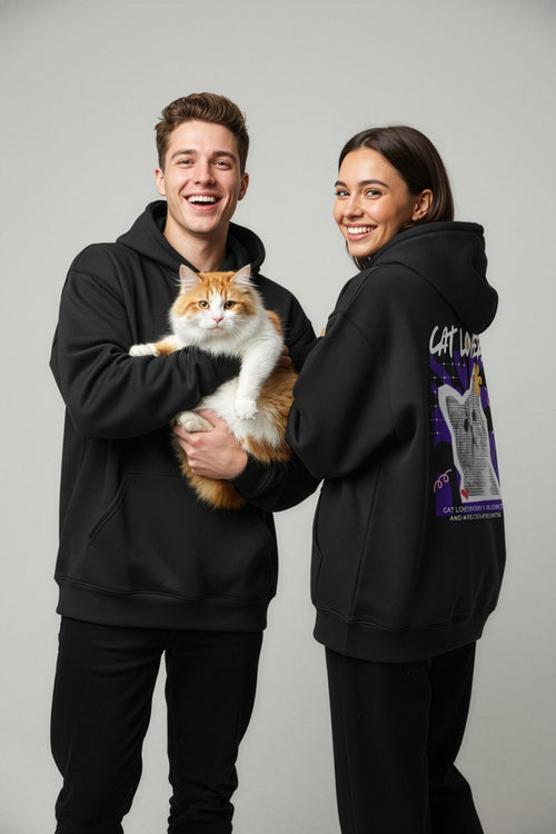 Two people wearing black hoodies, one holding a cat, on a plain background
