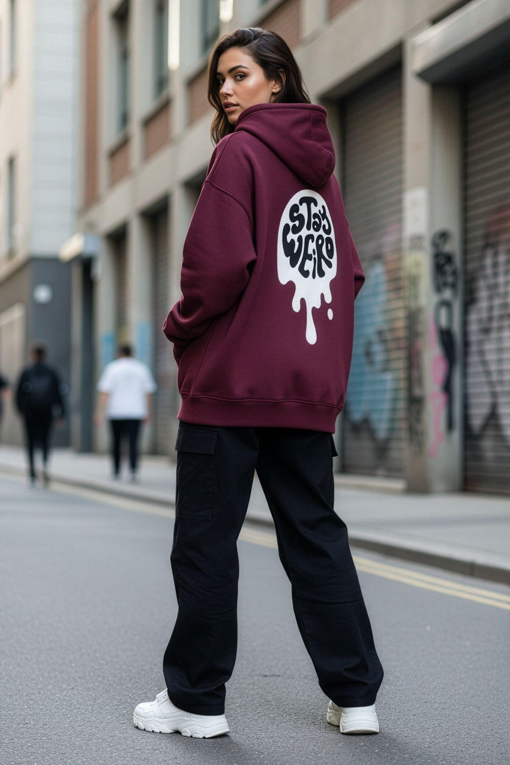 Person wearing a maroon hoodie with a logo on the back, standing on an urban street.
