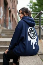Person wearing a navy hoodie with 'Stay Weird' text on the back, sitting on steps outdoors.