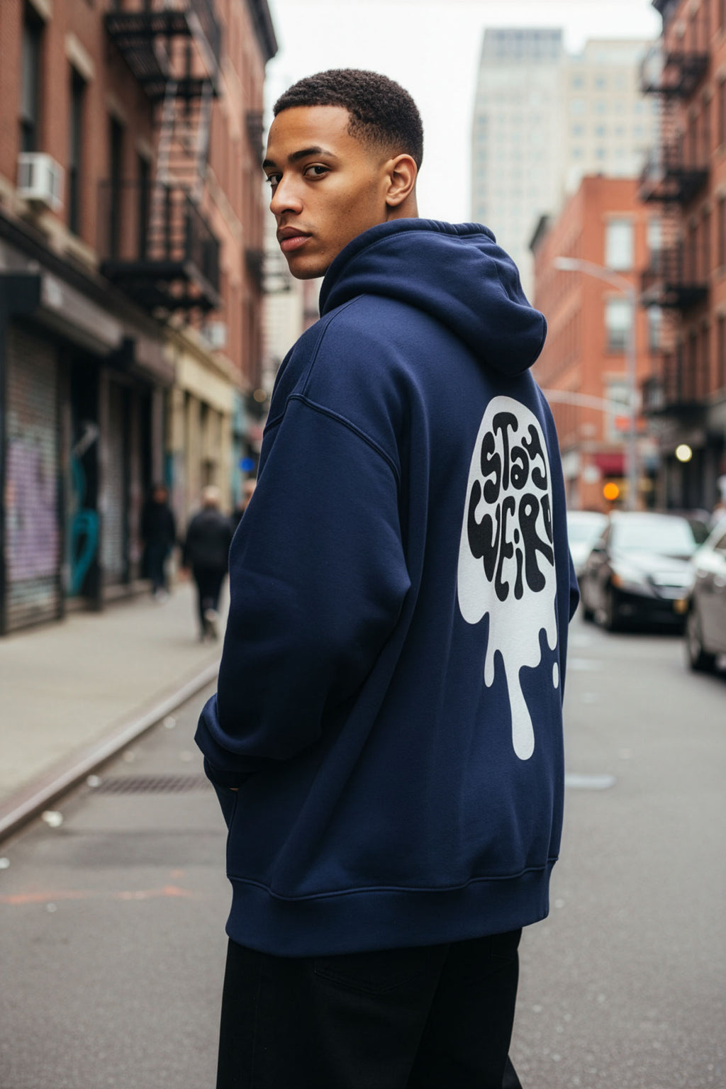 Person wearing a navy hoodie with a logo on the back, standing on a city street.