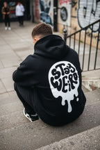 Person wearing a black hoodie with a white graphic design sitting on a street.