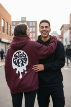 Two people hugging on a street with one wearing a maroon hoodie with 'Stay Weird' text.