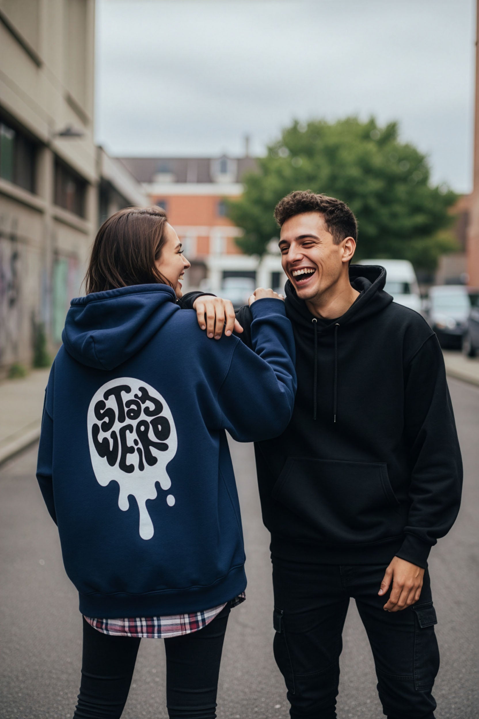 Two people standing on a street, one wearing a blue hoodie with a logo and the other in a black hoodie.